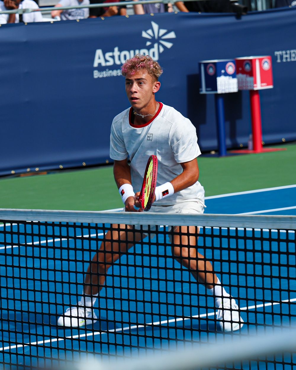 Young tennis player in white outfit holds racket at net on blue court with Astound Business Solutions signage.