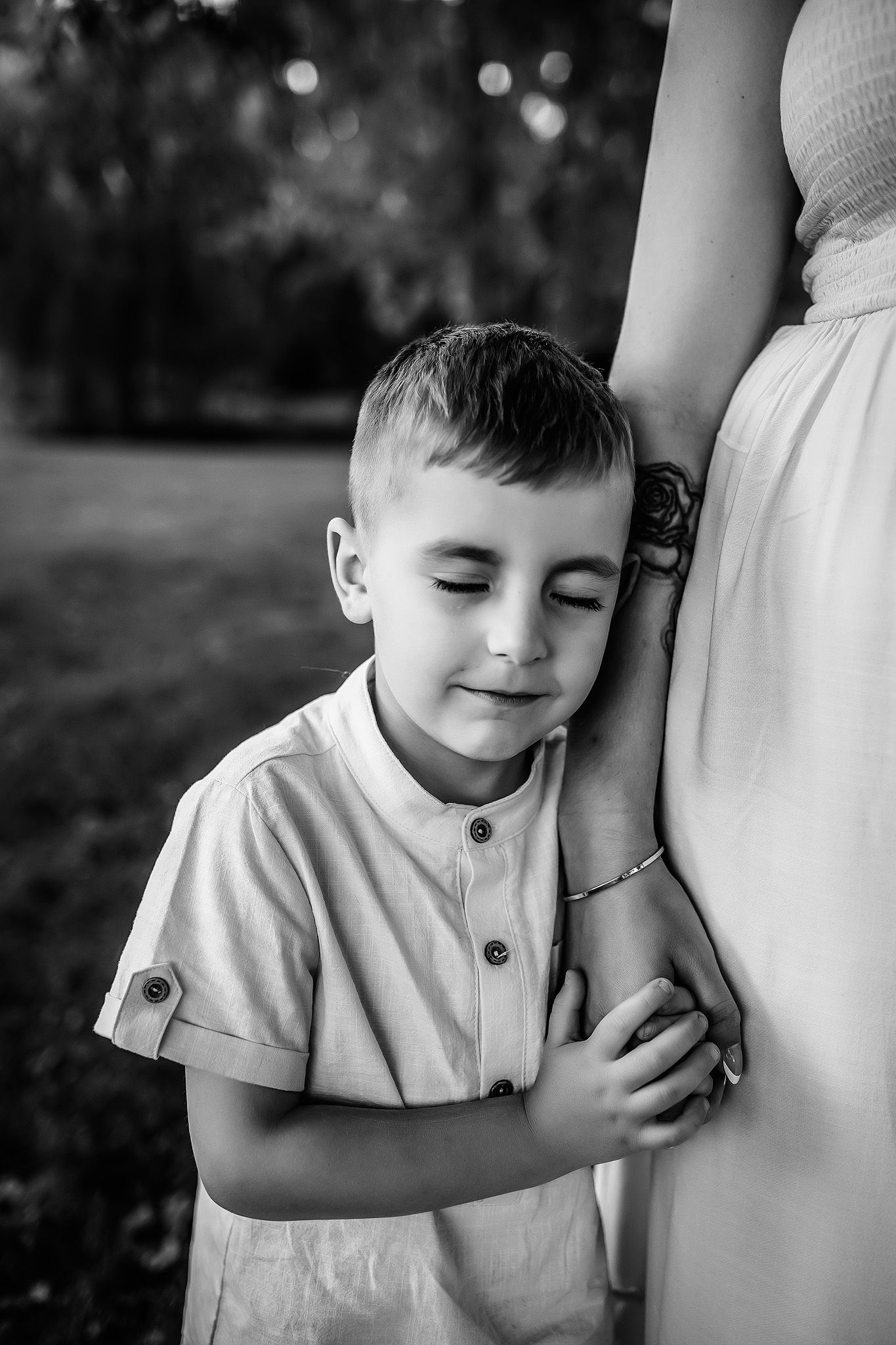 Black and white photo of a young boy leaning against his pregnant mother, smiling with eyes closed outdoors.