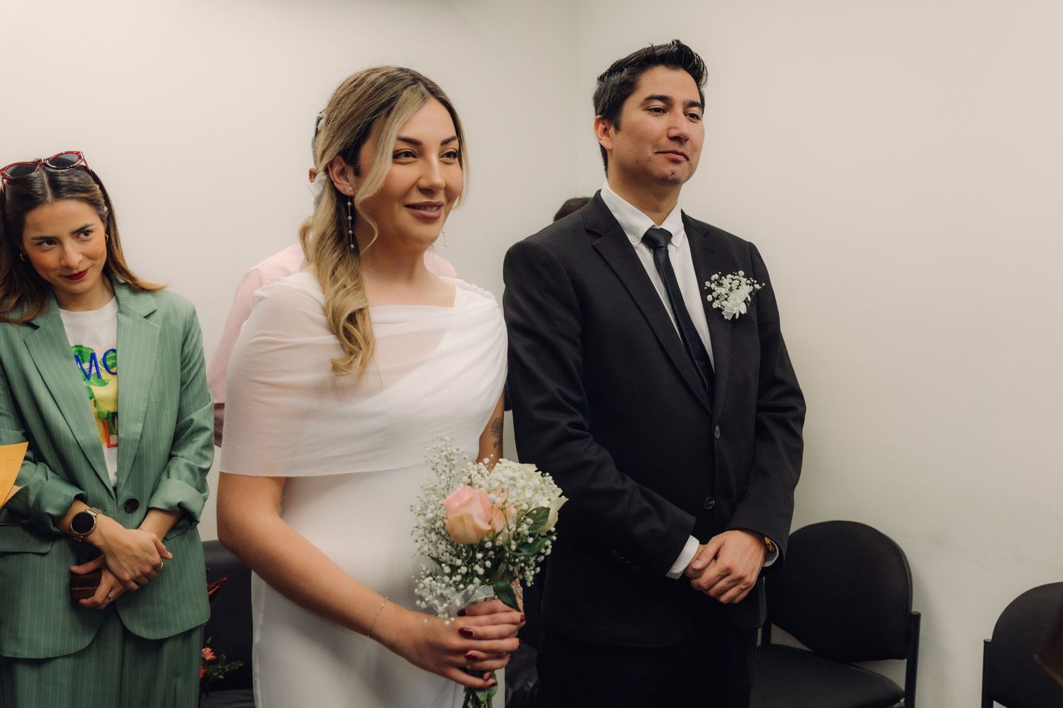 Smiling blonde bride holding bouquet stands beside groom in black suit during civil wedding ceremony indoors.