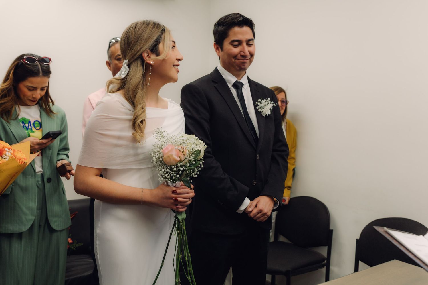 Bride in white cape dress laughing with groom at civil wedding ceremony, guests visible in background.