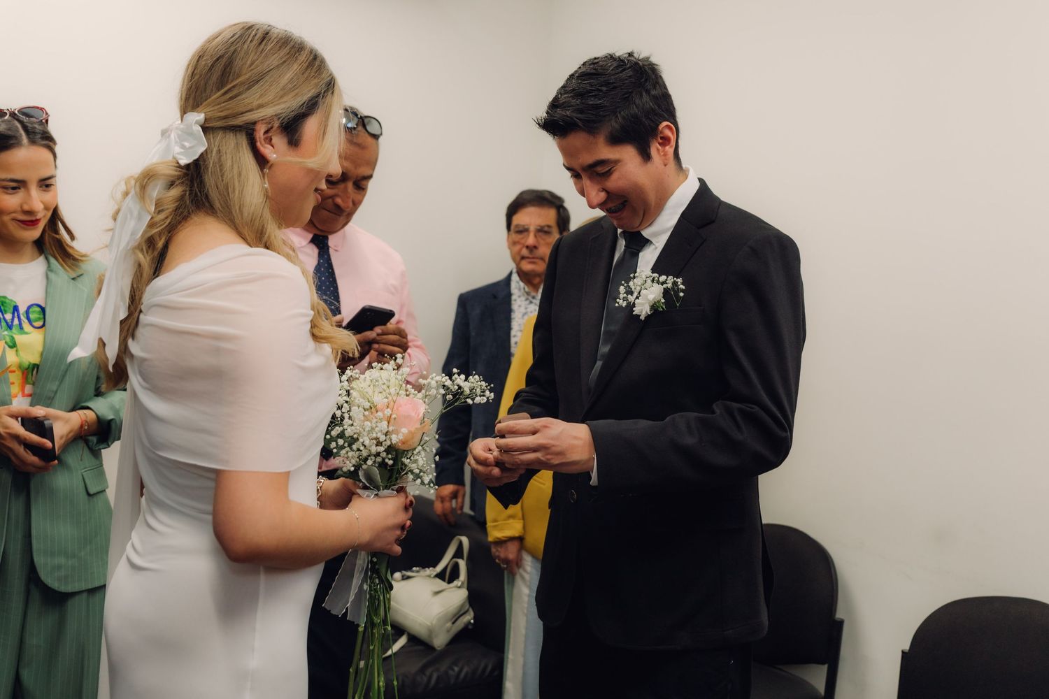 Groom in black suit placing wedding ring on bride's finger during intimate indoor civil ceremony.