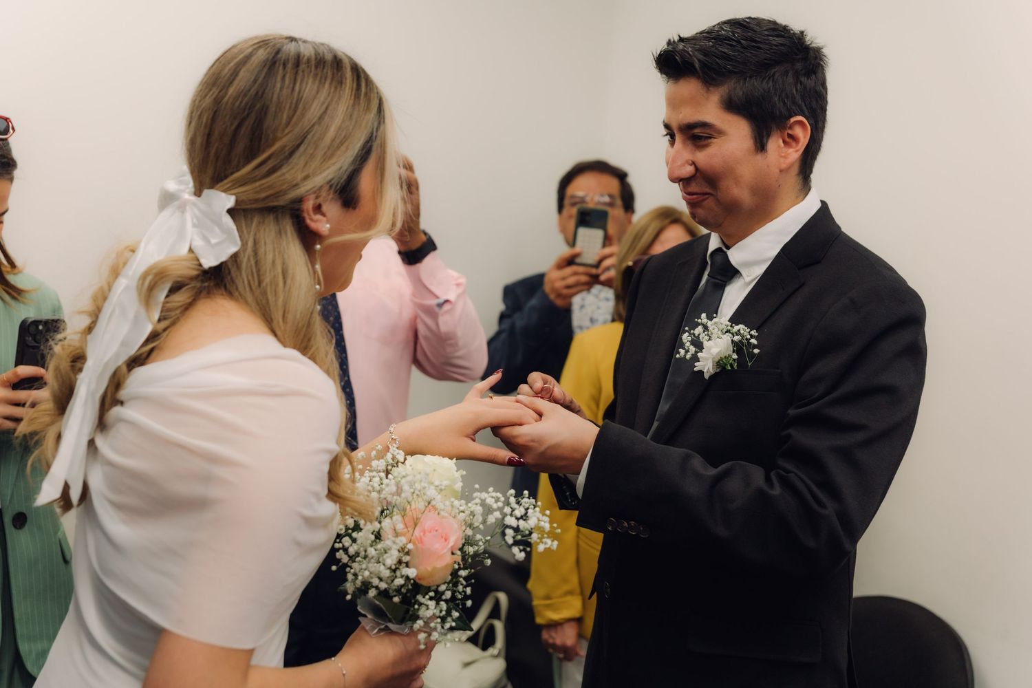 Couple exchanging wedding rings during small civil ceremony with guests capturing the moment on phones.