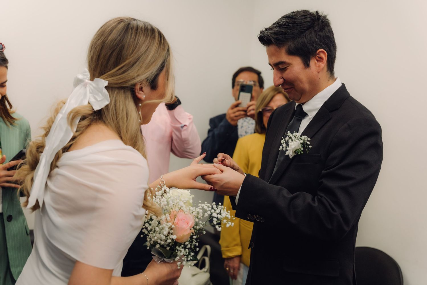 Groom placing wedding ring on bride's finger during intimate civil ceremony while guests watch and photograph.