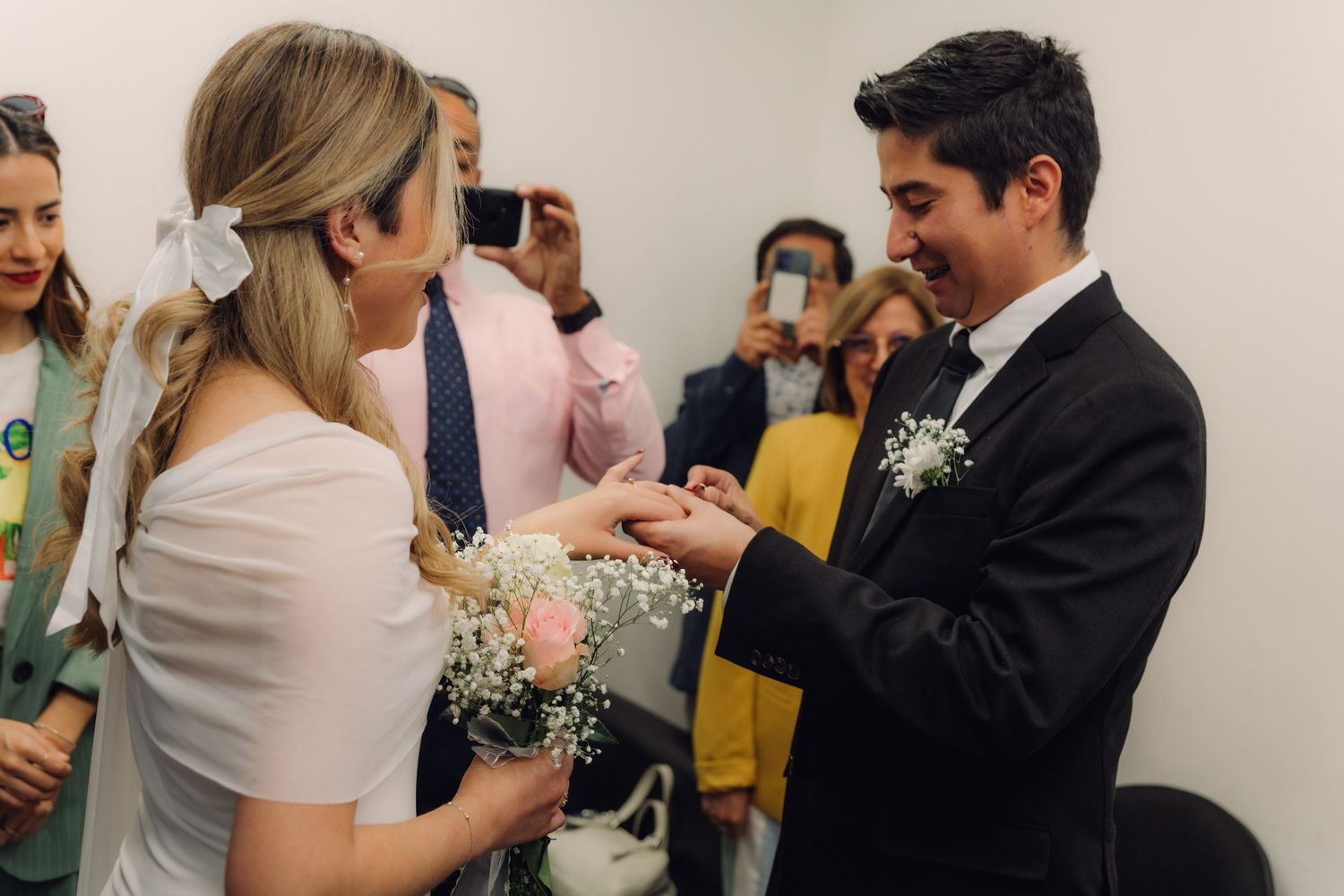 Smiling groom sliding wedding band onto bride's finger as wedding guests witness the ring exchange.
