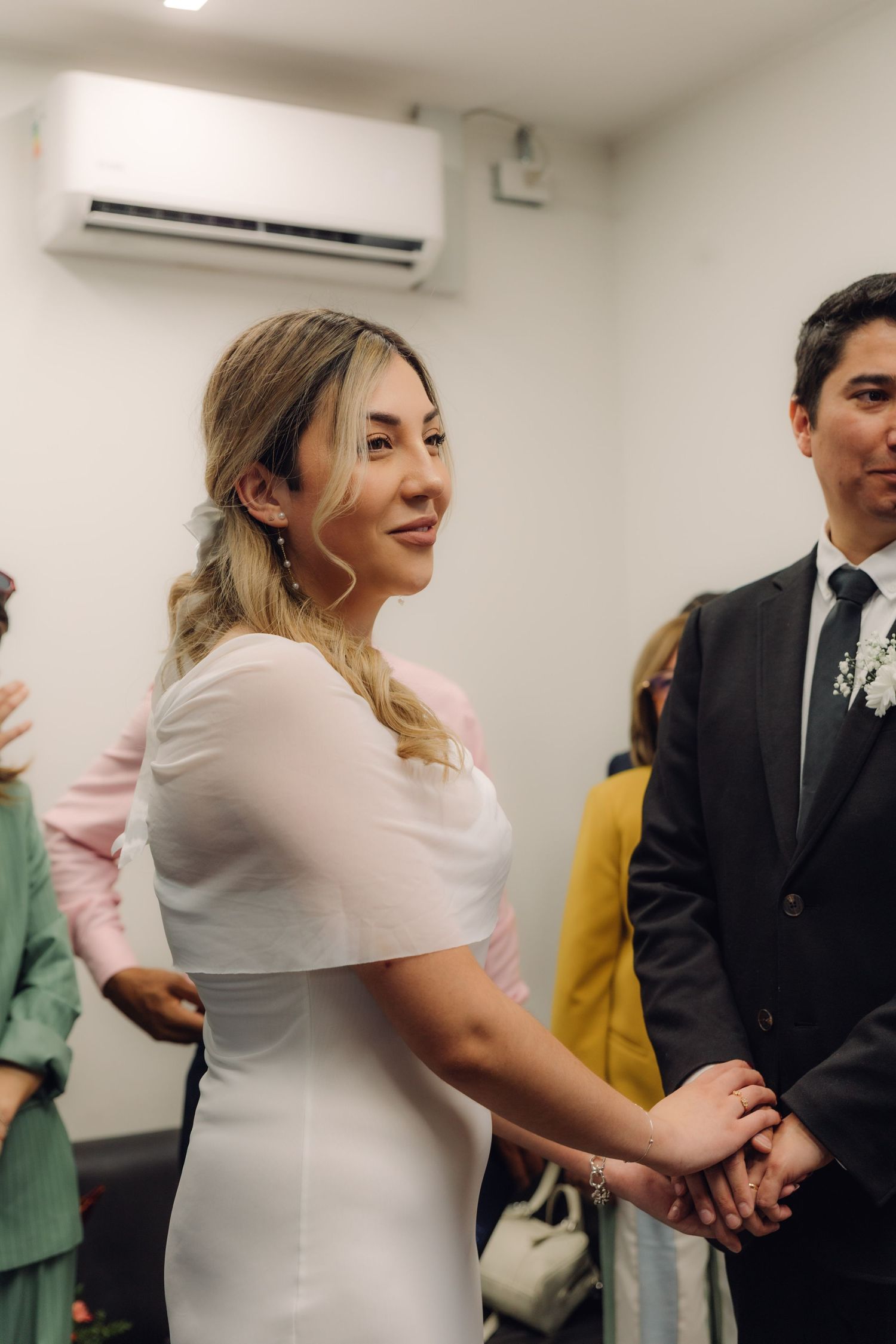 Blonde bride in white dress holding groom's hands and smiling during civil wedding ceremony indoors.