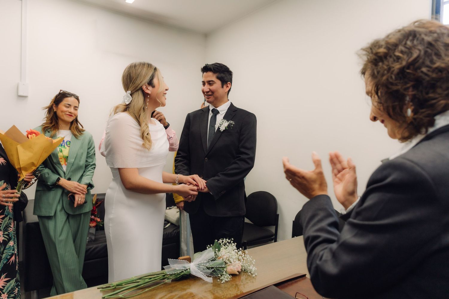 Newlyweds smiling at each other as wedding guest applauds during small civil marriage ceremony.