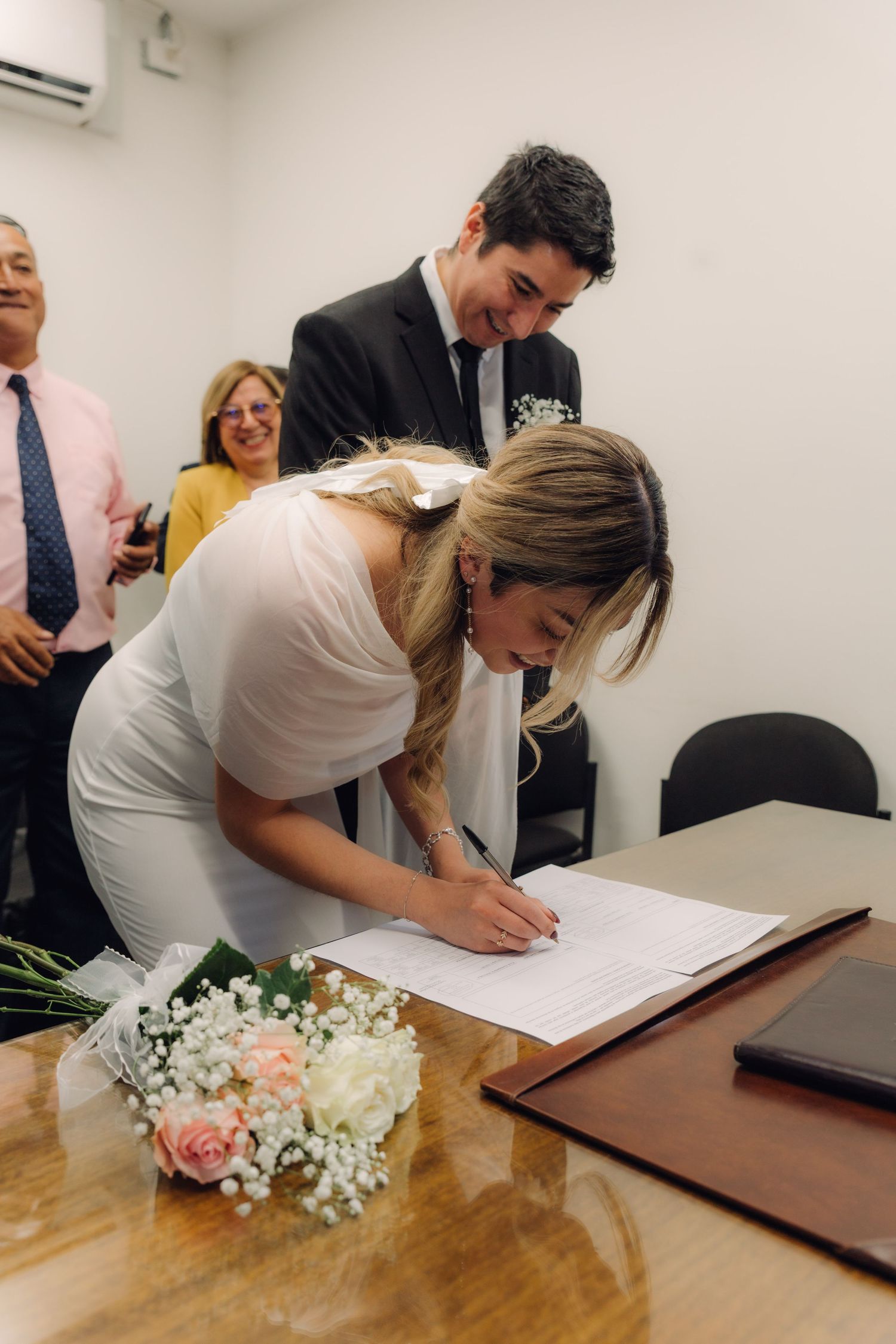 Bride in white dress signing official marriage documents at wooden table with groom watching.