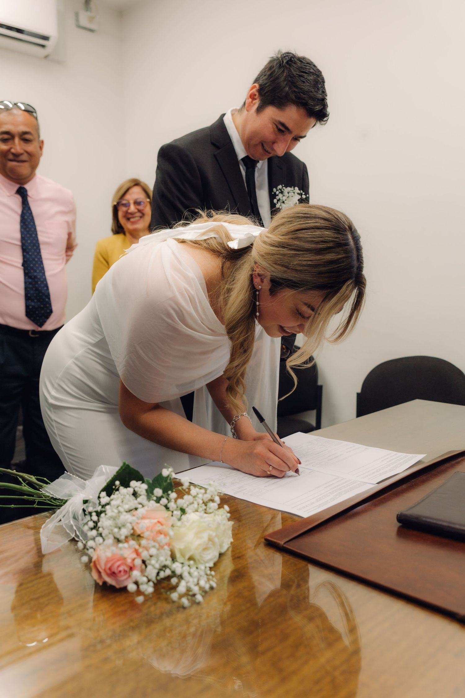 Blonde bride signing wedding registry documents at wooden table with groom and family watching.