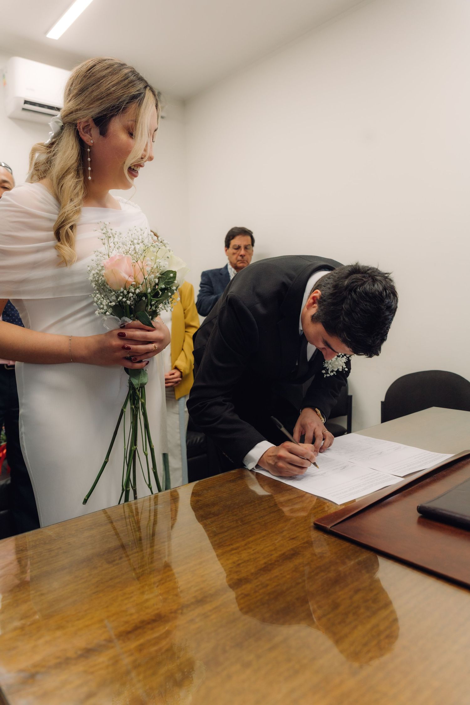 Groom signing official marriage certificate at wooden table while bride holds bouquet and smiles.