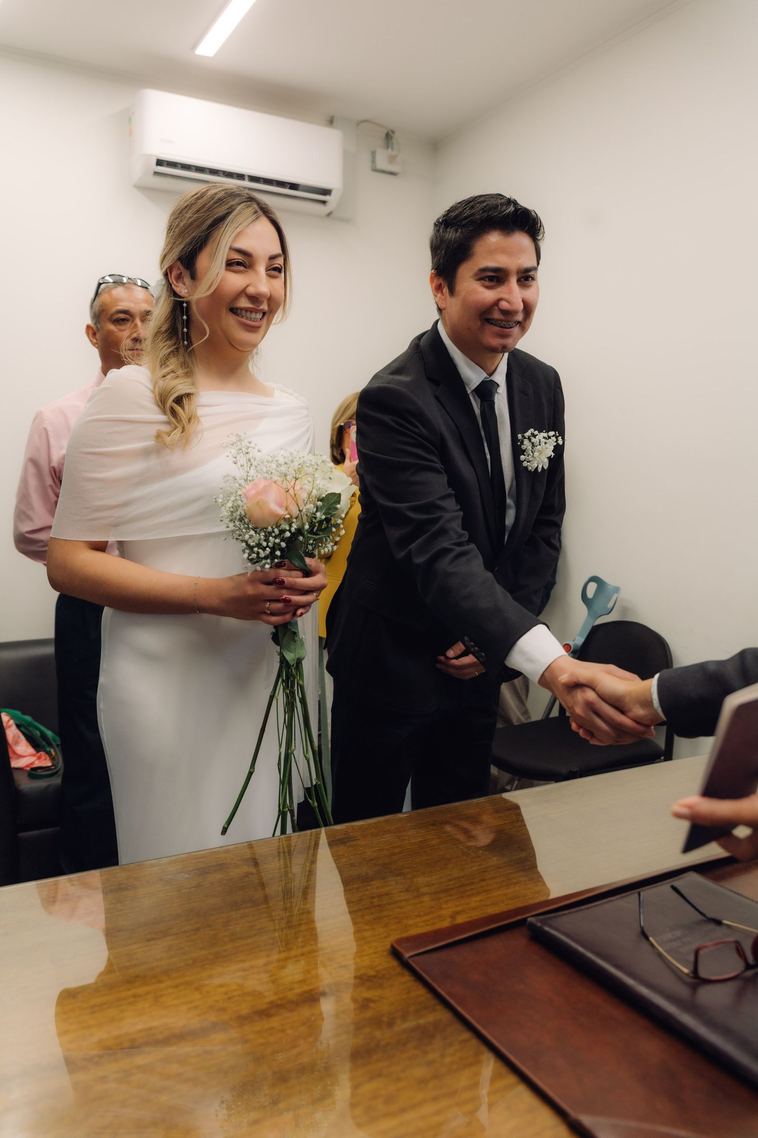 Smiling bride holding pink bouquet and groom shaking hands during civil wedding ceremony indoors.