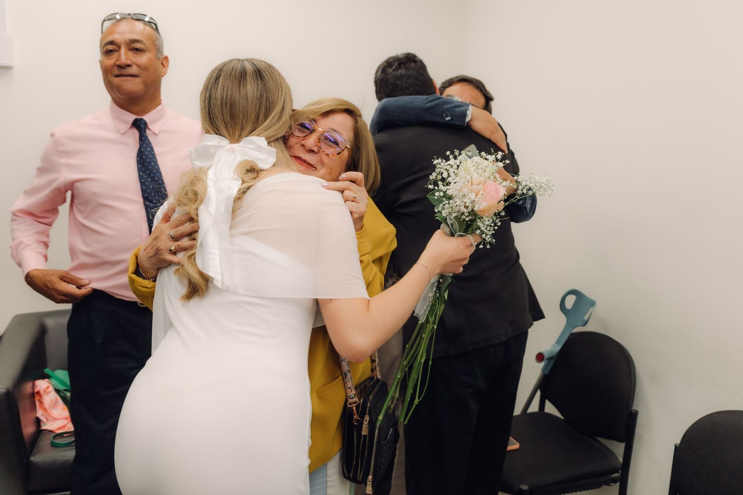 Newlywed bride embracing a woman in yellow jacket during post-wedding congratulations in office setting.