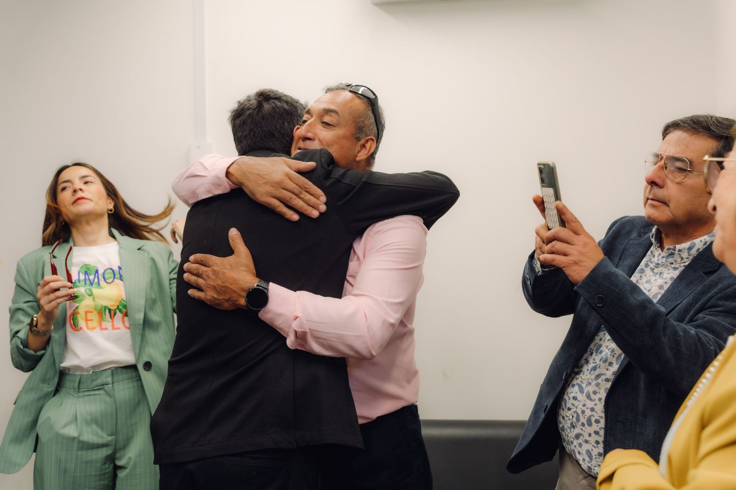 Two men sharing a warm hug while guests photograph the moment at a wedding celebration.