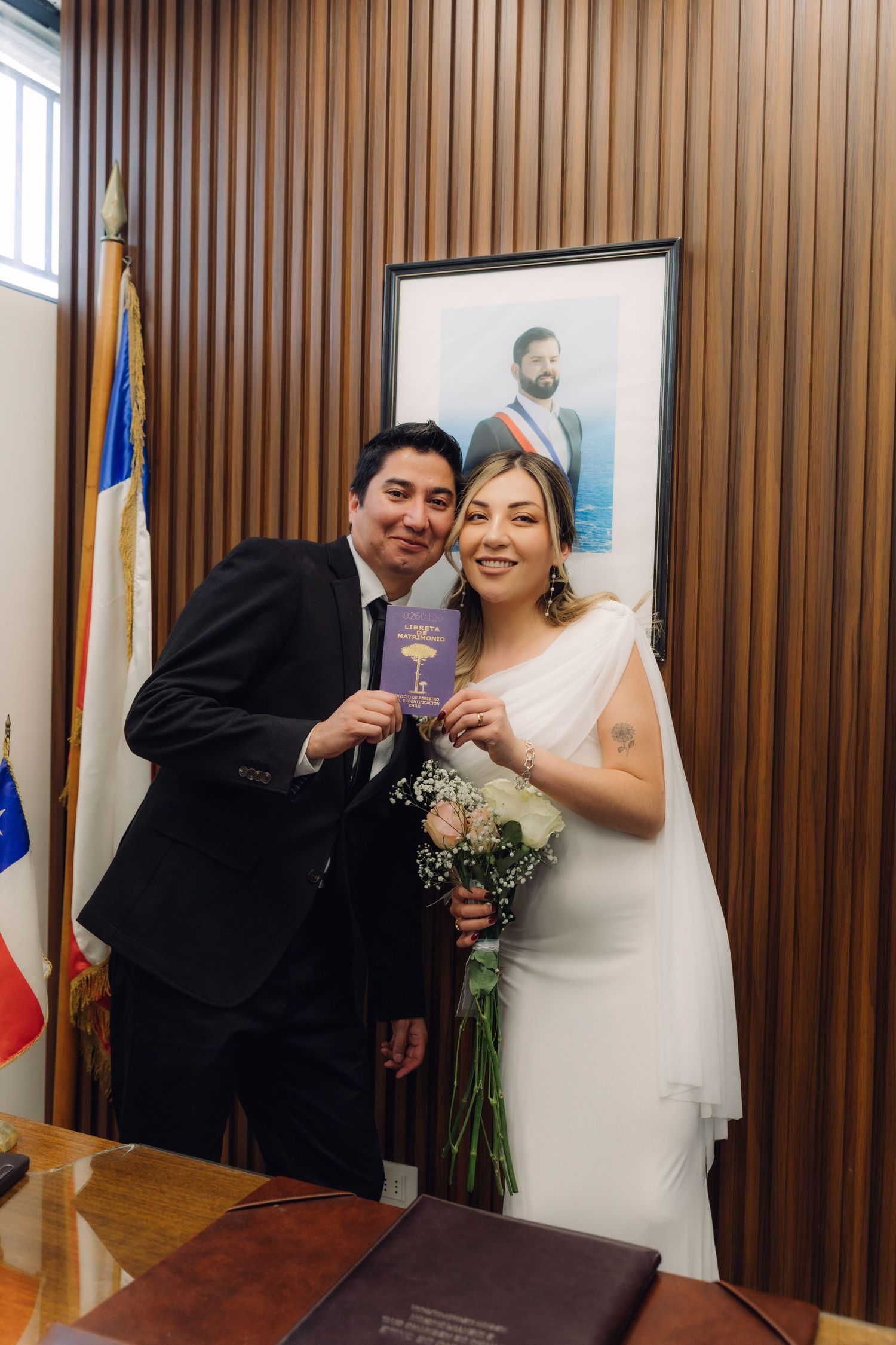 Newlyweds posing with marriage certificate in front of a portrait and Chilean flag after civil ceremony.