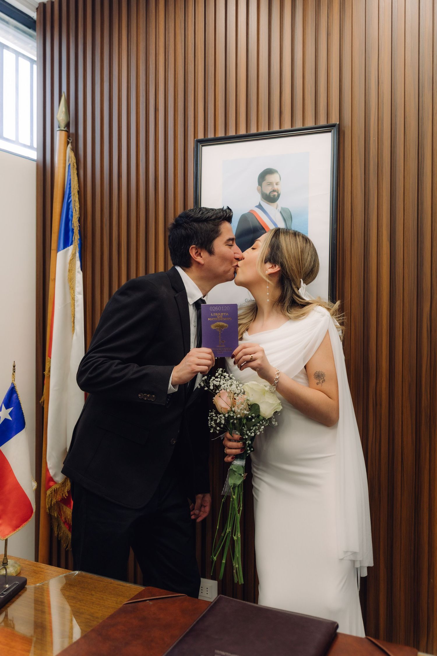 Bride and groom sharing first kiss holding marriage booklet at Chilean civil registry office.