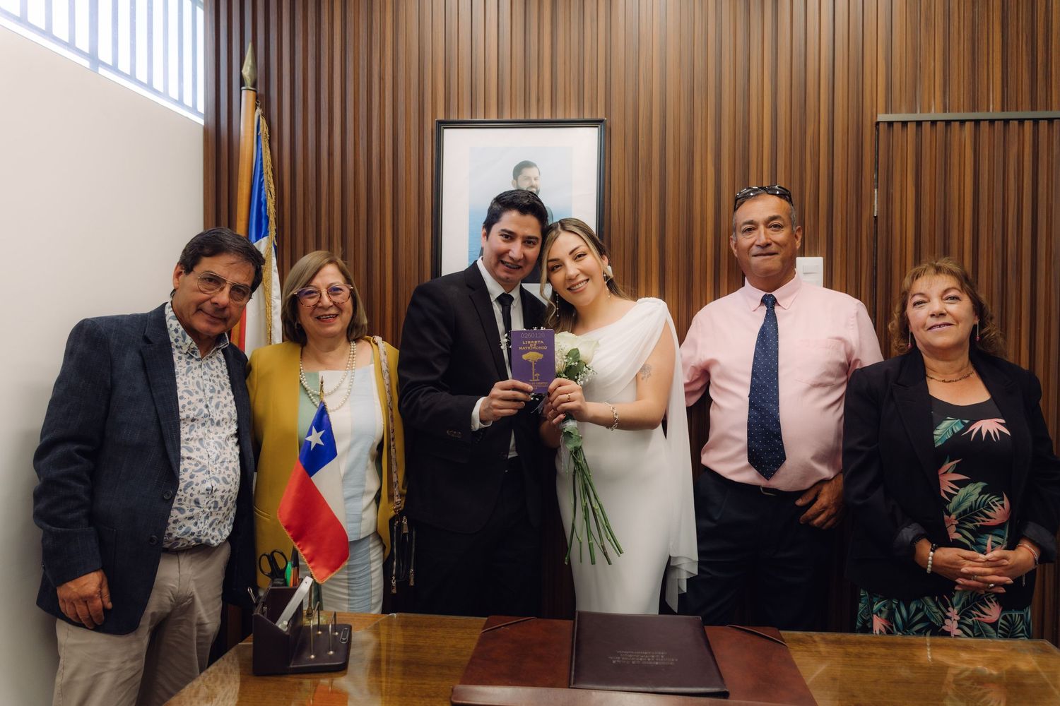 Newlyweds posing with six family members at Chilean civil wedding ceremony with flag backdrop.