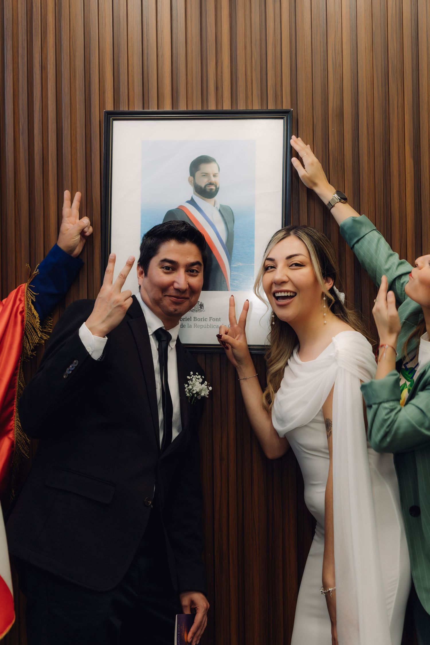 Laughing newlyweds making peace signs in front of a framed portrait with celebrating guests around them.
