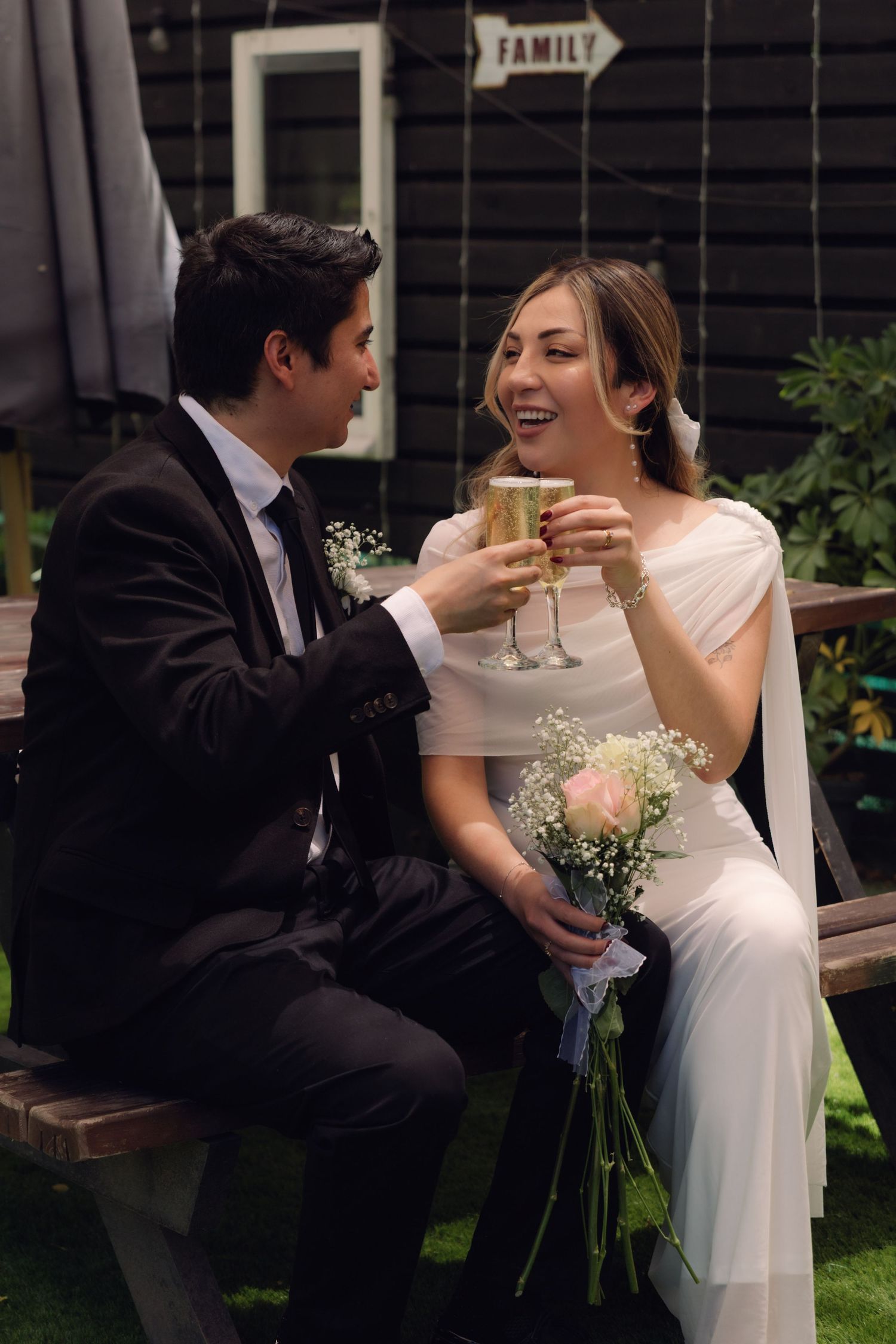Bride and groom toasting with champagne glasses while sitting on a bench, bride holding a pink rose bouquet.