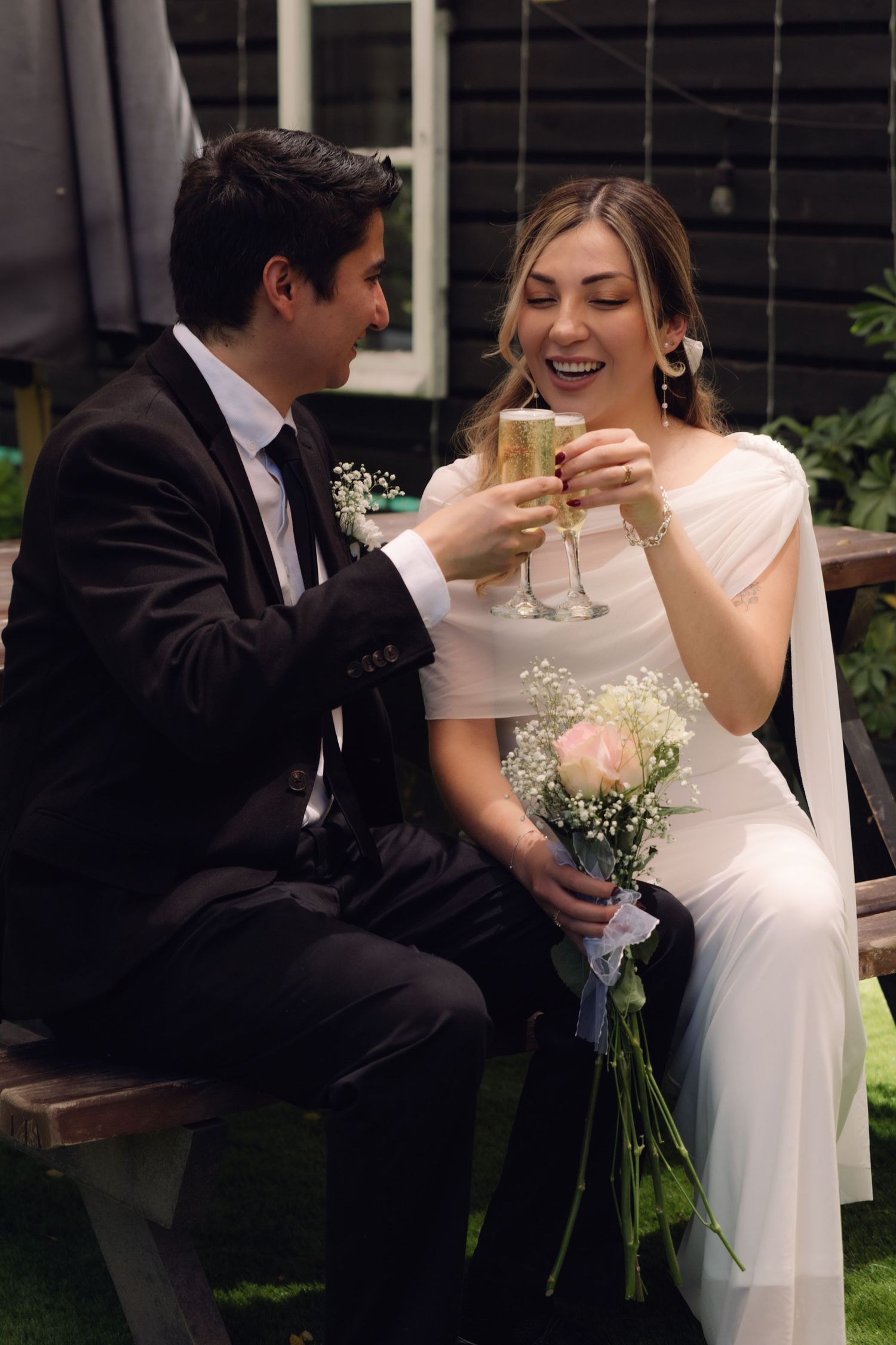 Happy newlyweds clinking champagne flutes outdoors, bride in white dress holding a pink rose bouquet.