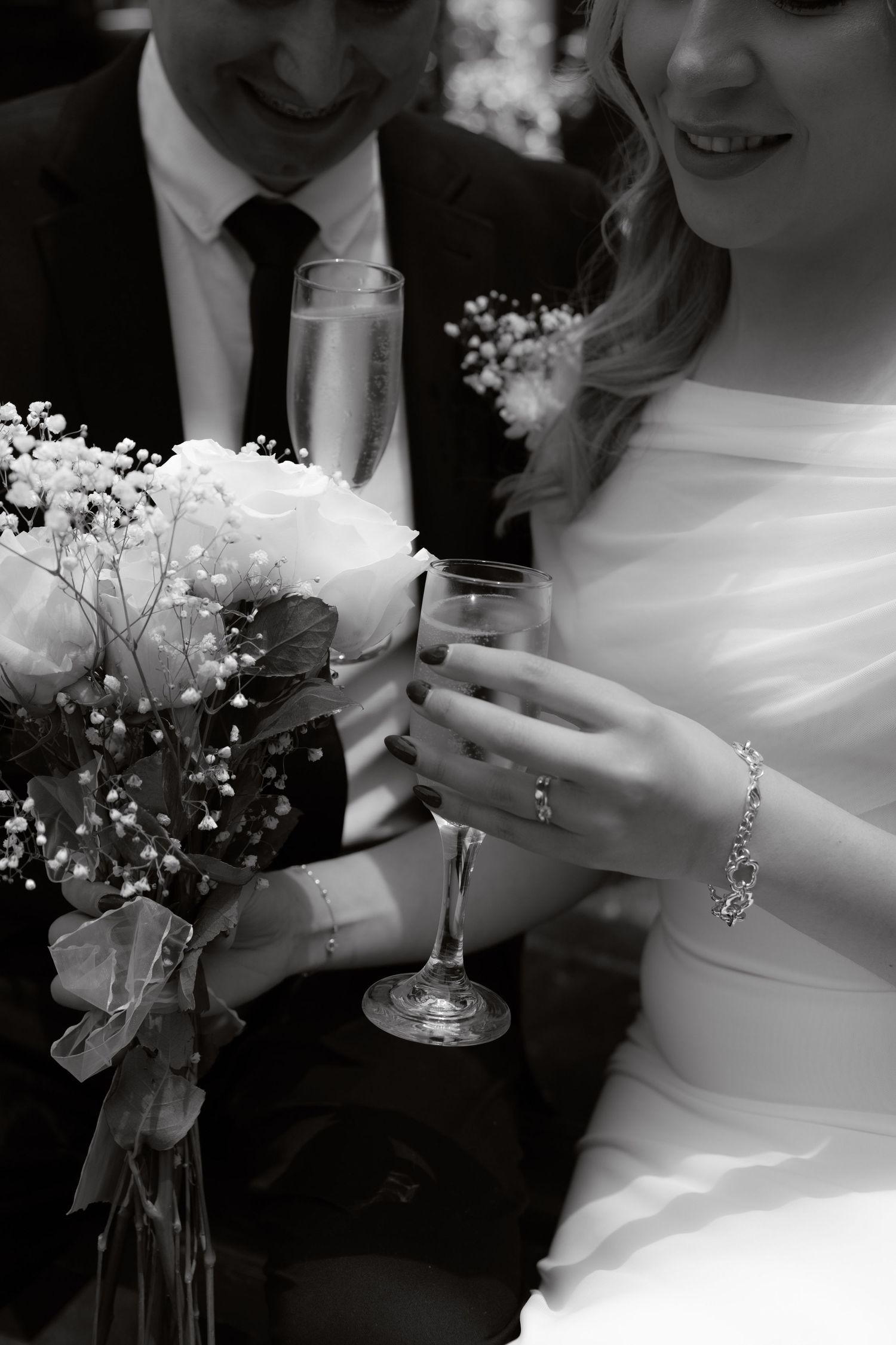 Black and white photo of newlyweds holding champagne flutes with a bridal bouquet of roses.