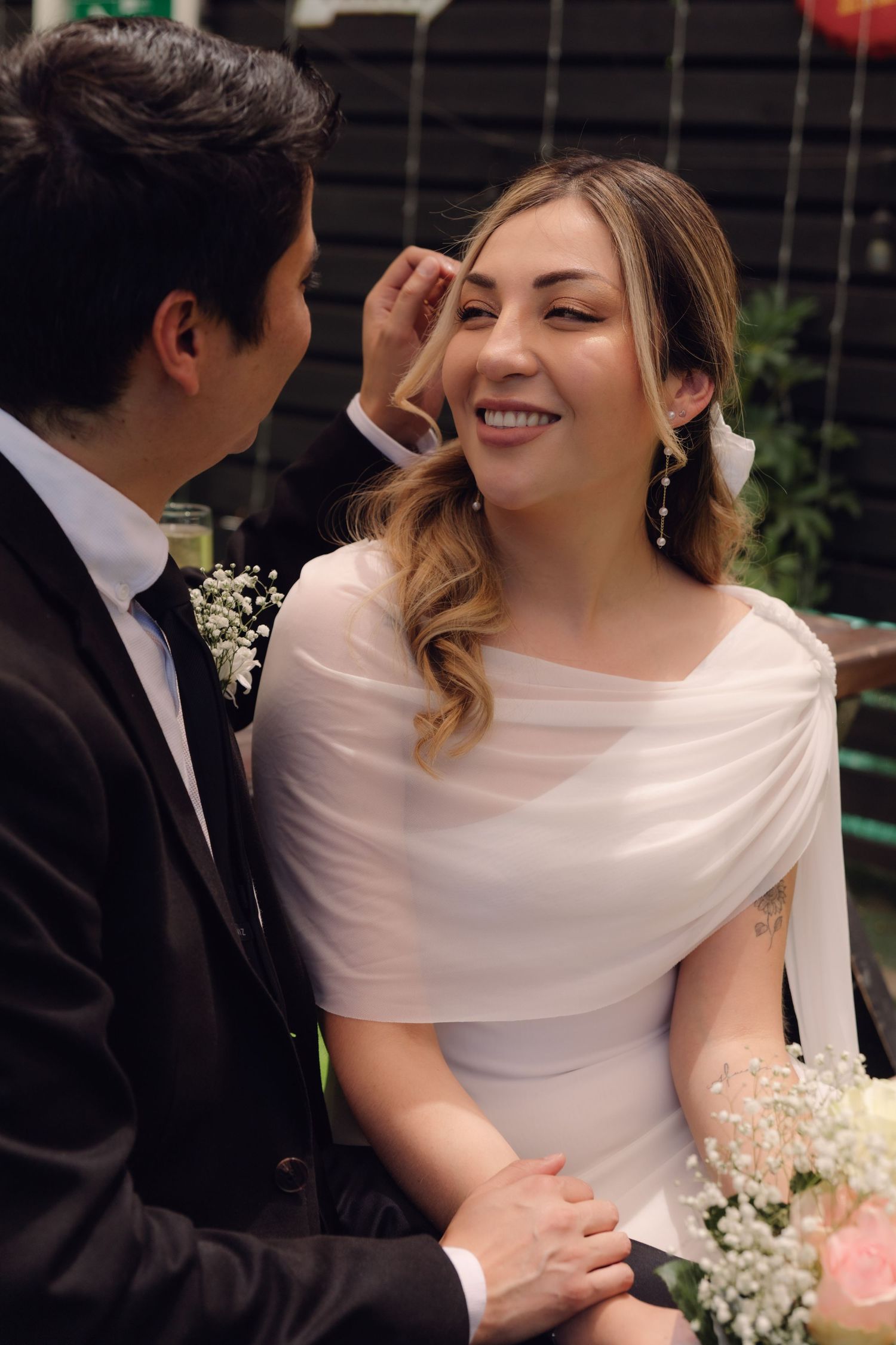 Groom gently brushing bride's hair as she smiles warmly, bride wearing drop earrings and white dress.