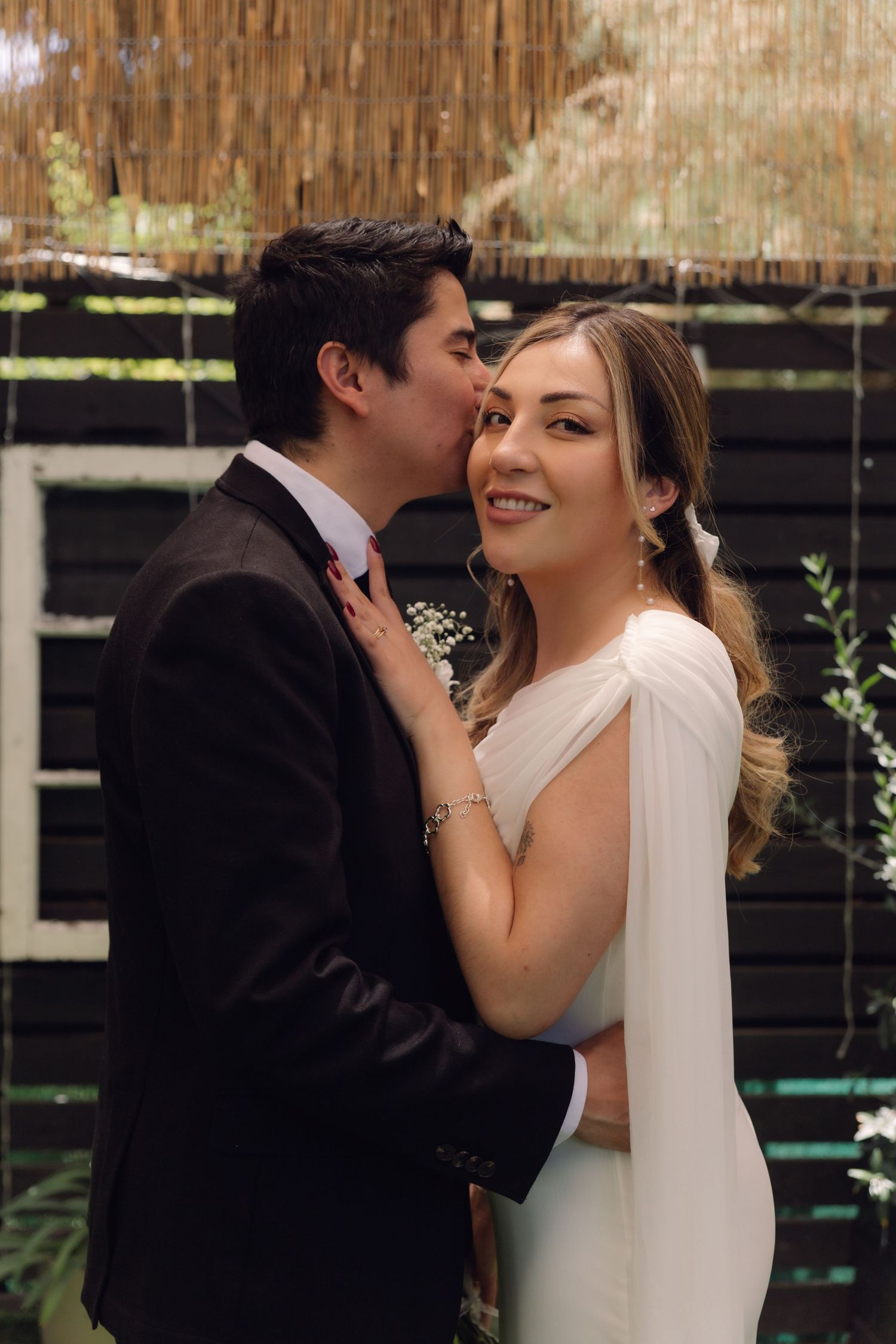 Groom kissing bride on the cheek as she smiles at camera, standing in front of a dark wooden backdrop.