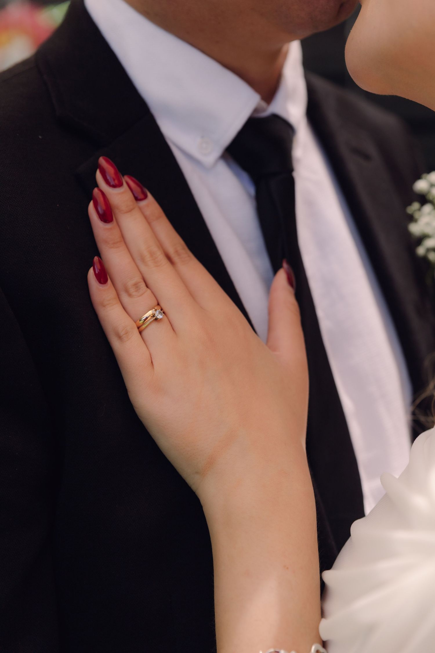 Close-up of bride's hand with gold wedding ring and red nails resting on groom's black suit jacket.