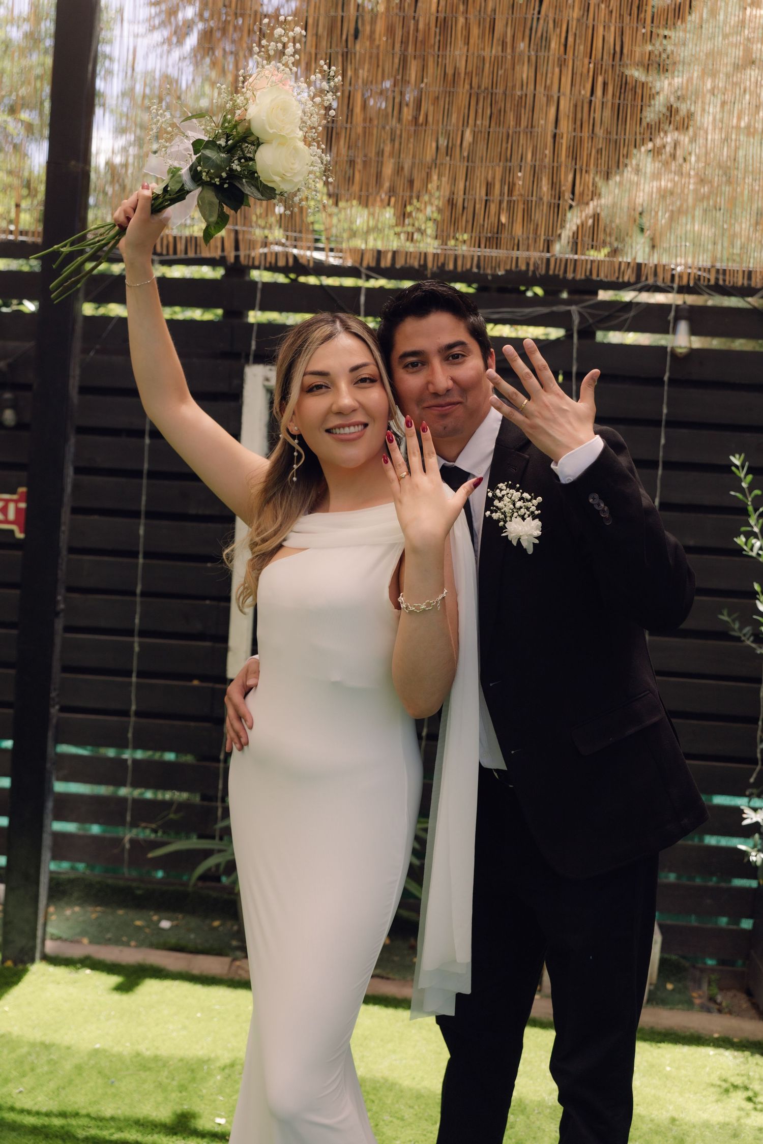 Joyful newlyweds showing wedding rings, bride in white gown holding bouquet aloft, groom in black suit, outdoor ceremony.