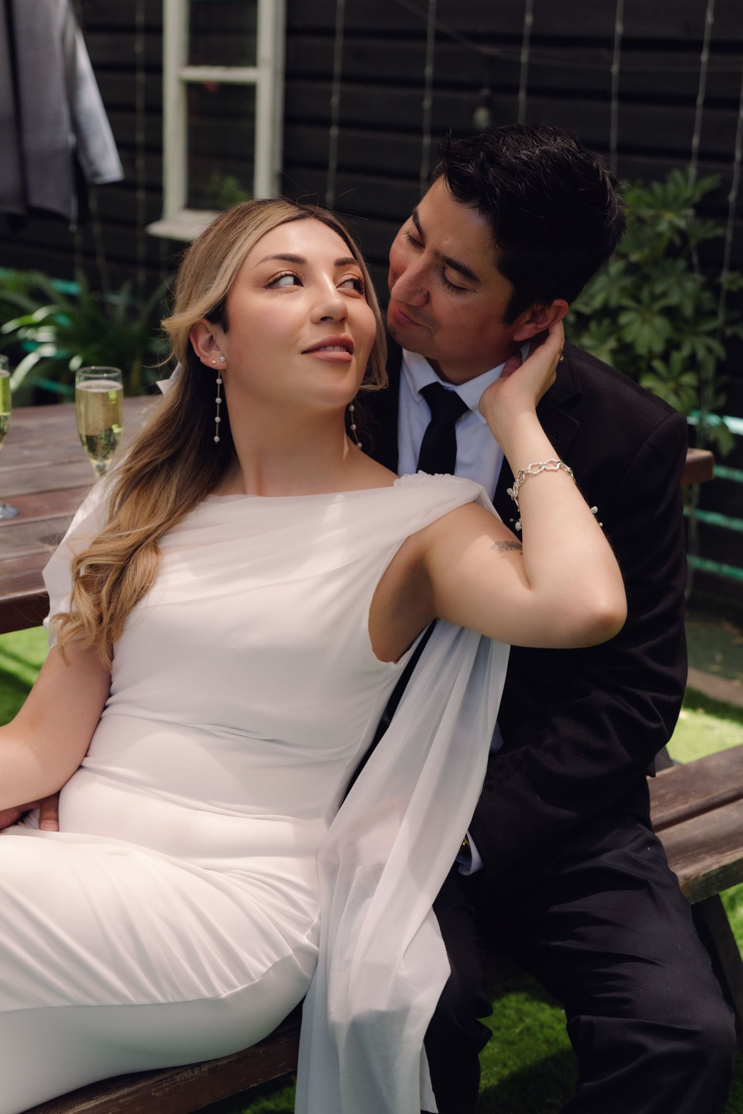 Groom in black suit kissing bride's cheek as she sits smiling, outdoor wedding reception with champagne.