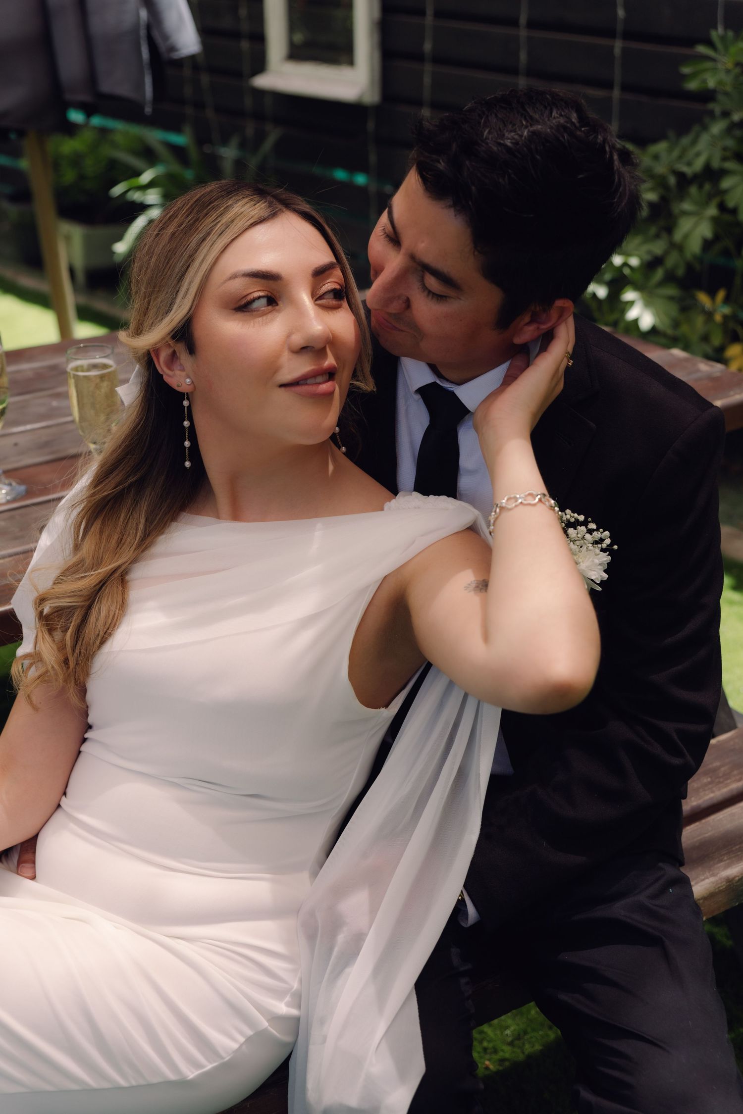 Romantic wedding moment with groom kissing bride's cheek, bride in white gown, seated outdoors at reception.