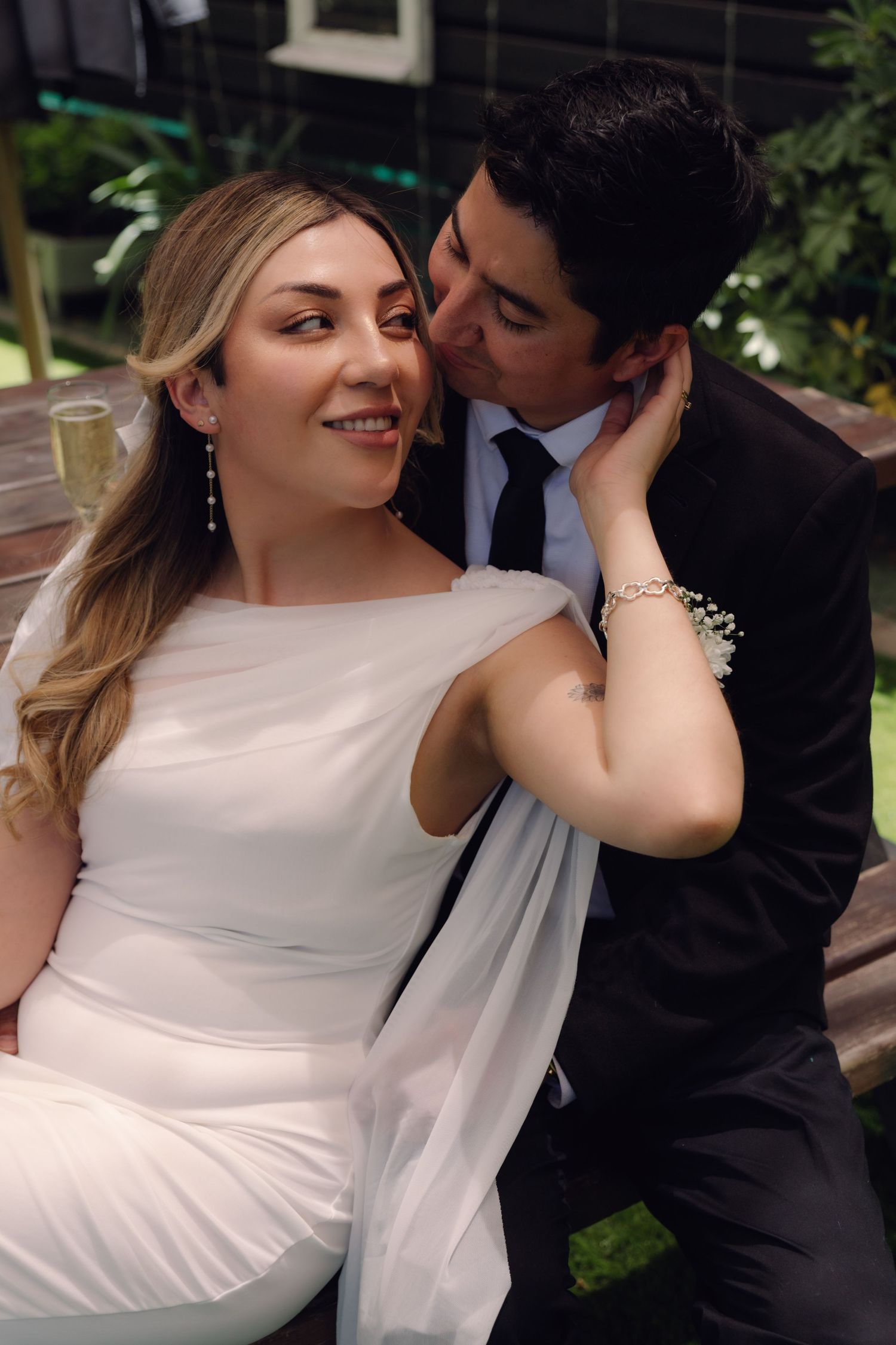 Groom tenderly kissing bride's cheek outdoors, bride in white gown smiling, wedding reception garden setting.