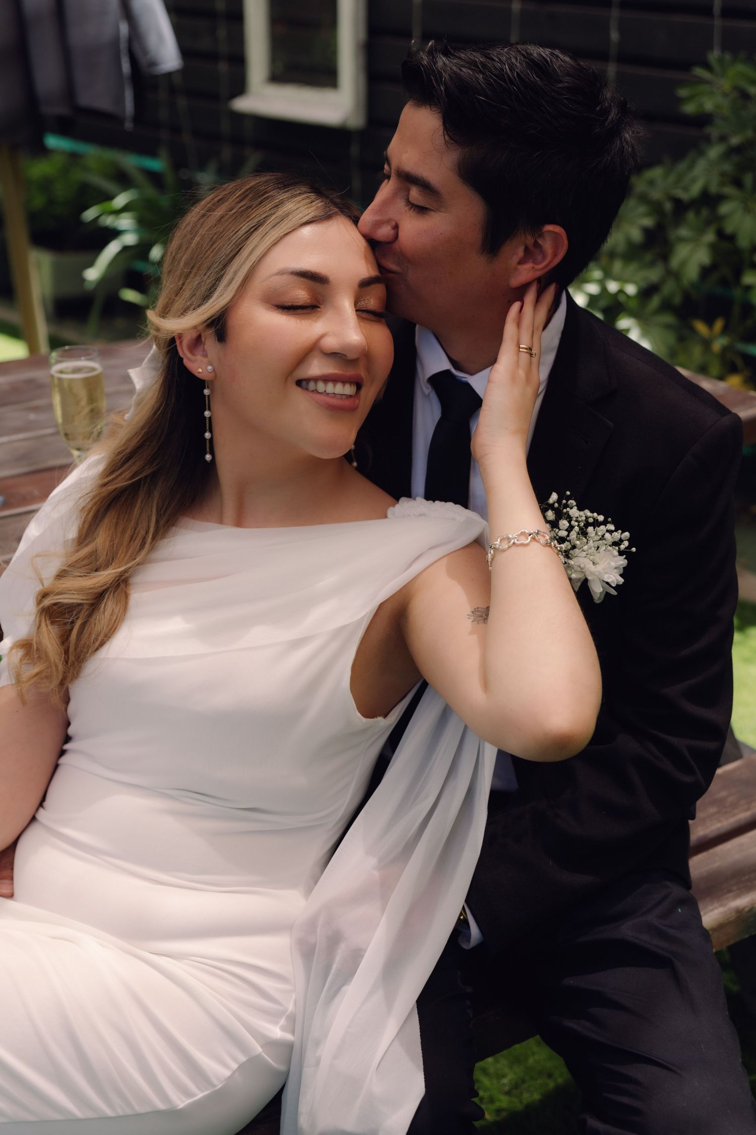 Groom kissing smiling bride on cheek at outdoor wedding reception, bride in white gown holding champagne.