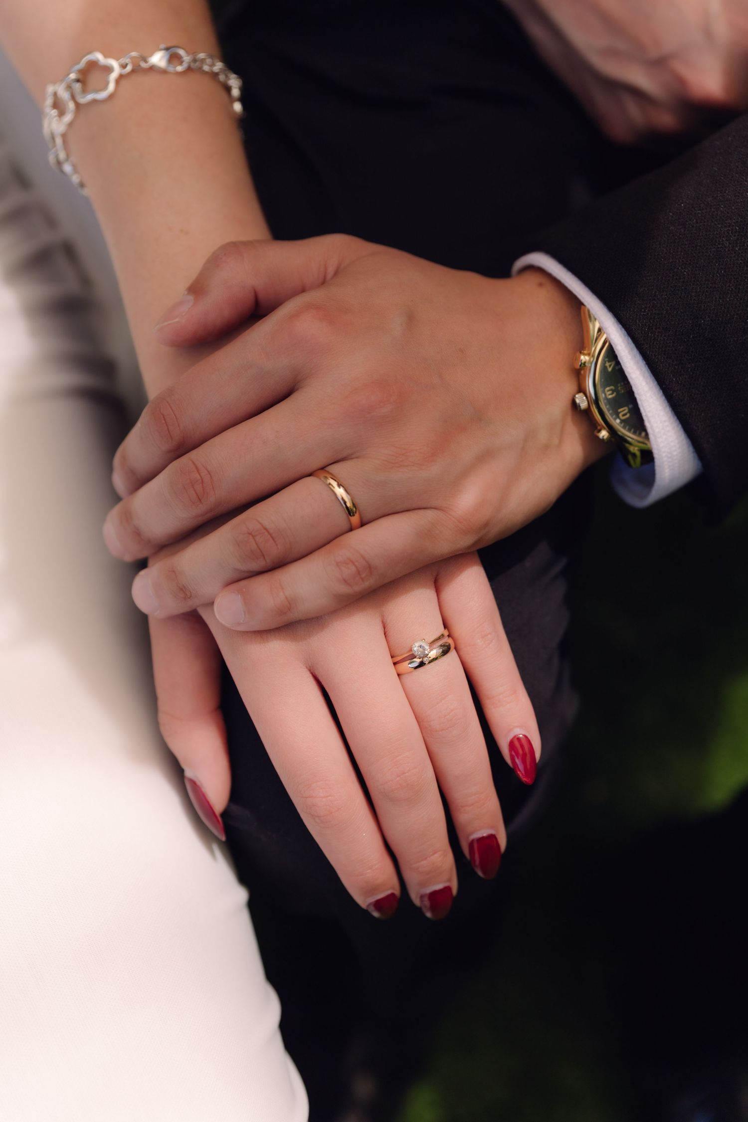 Close-up of newlyweds hands showing gold wedding rings, bride with red nails and bracelet, groom with watch.