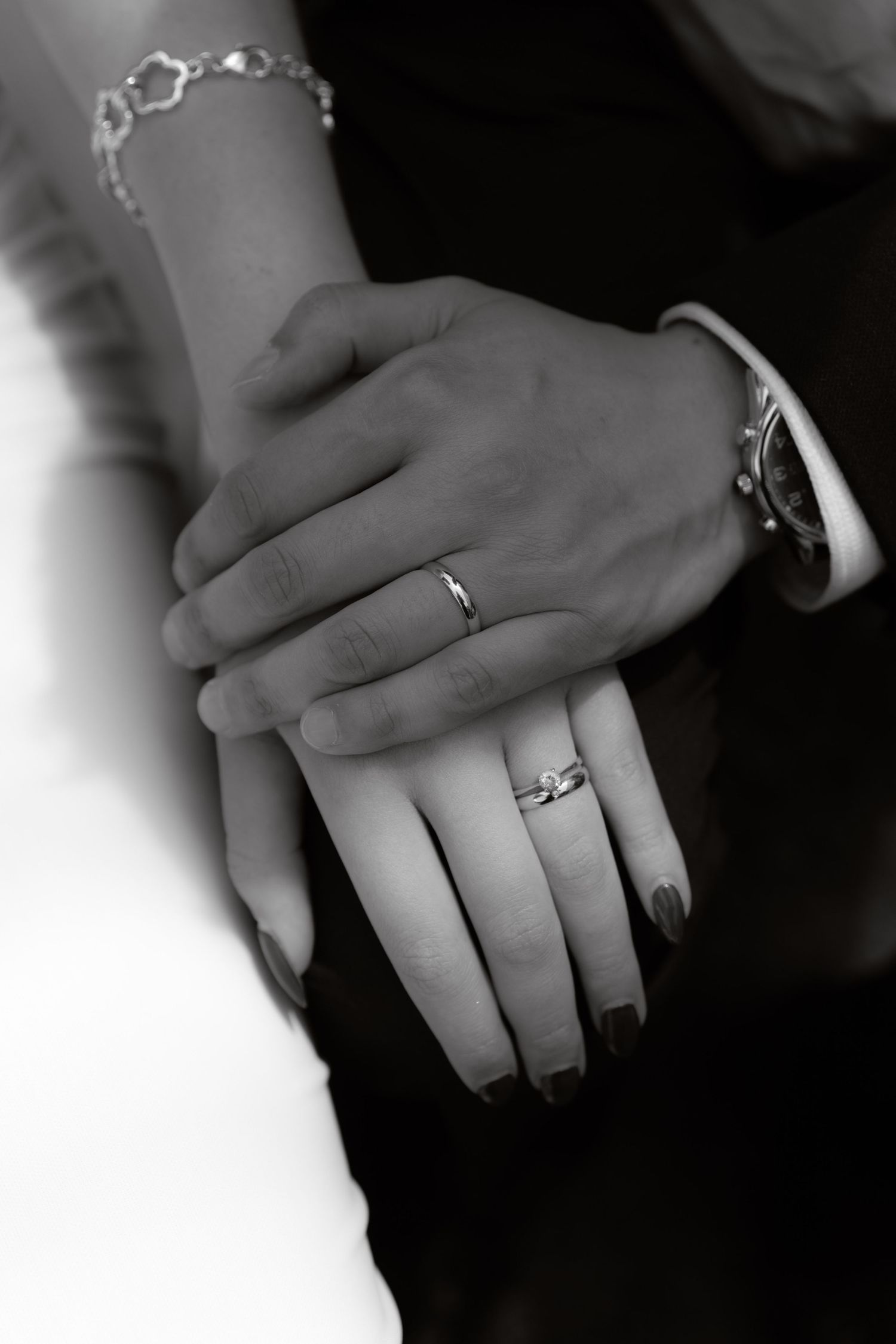 Black and white close-up of newlyweds hands displaying wedding rings, bride wearing bracelet, groom with watch.