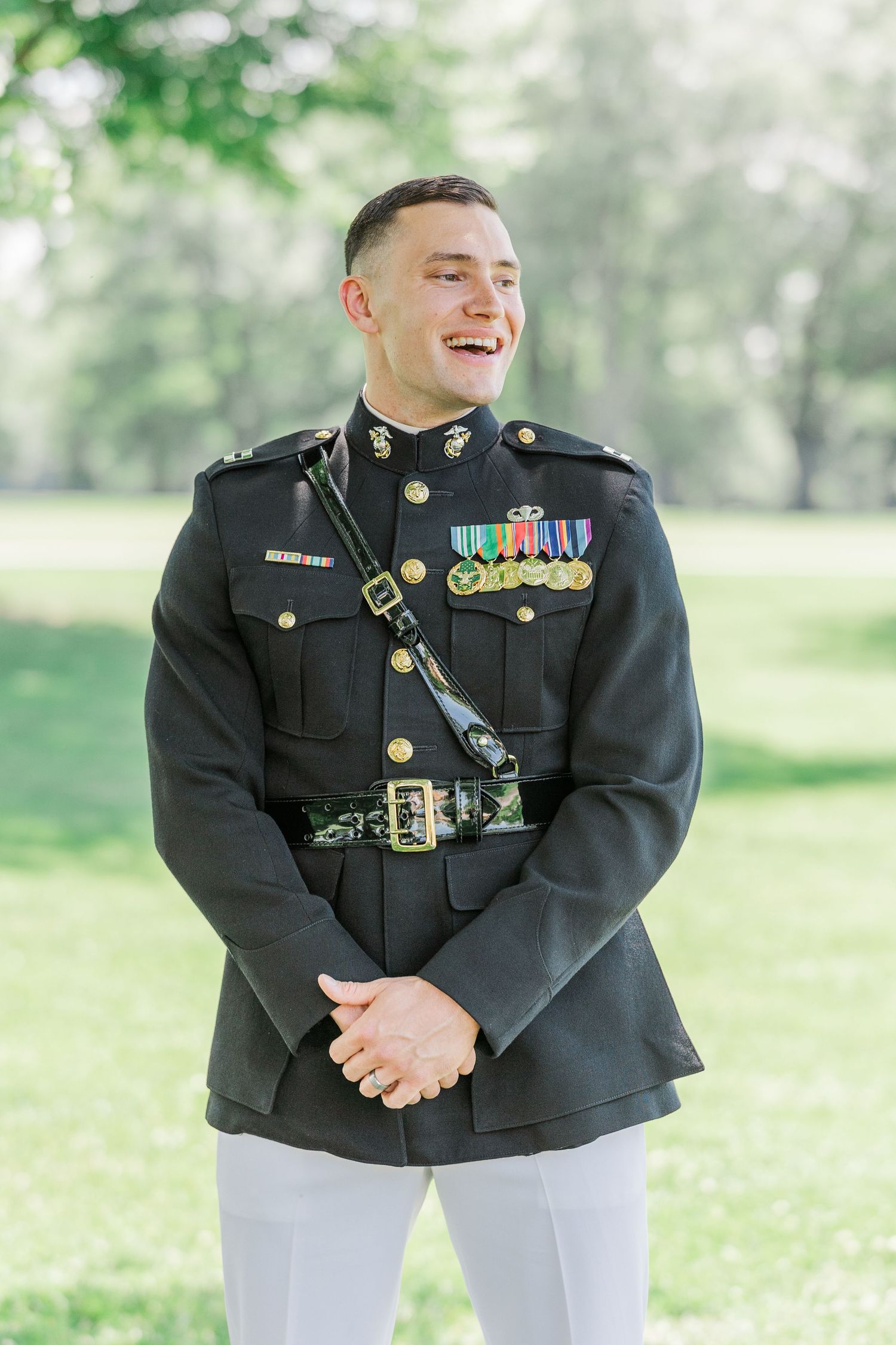 Smiling Marine in dress blues uniform with medals stands outdoors in a green park setting.
