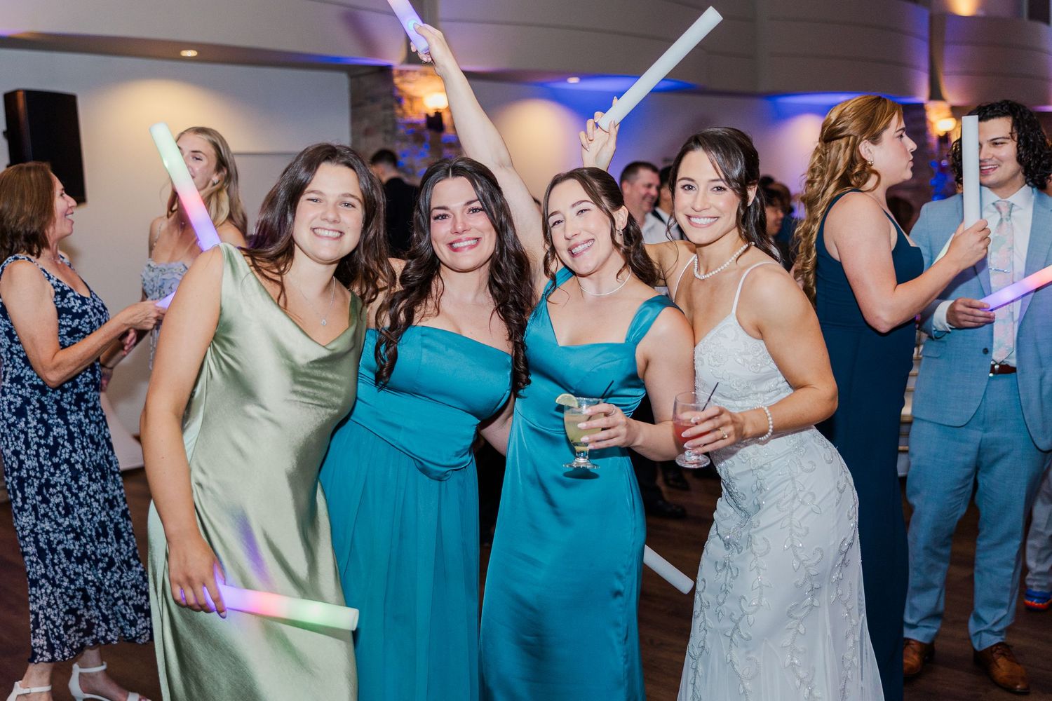 Four bridesmaids in teal and green gowns smile and dance with glow sticks at a wedding reception.