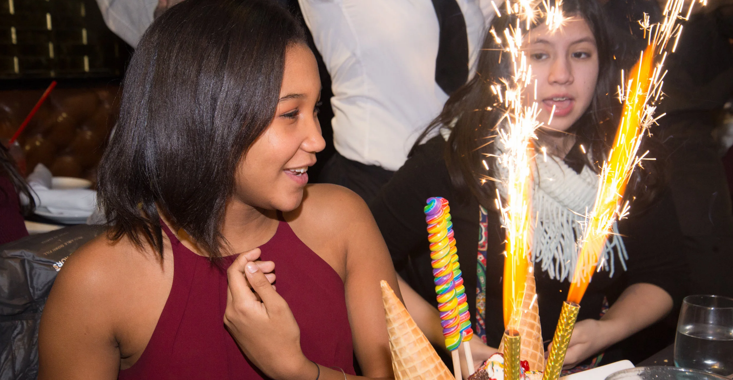 Two women celebrate a birthday with sparklers and colorful party decorations at a festive indoor event.