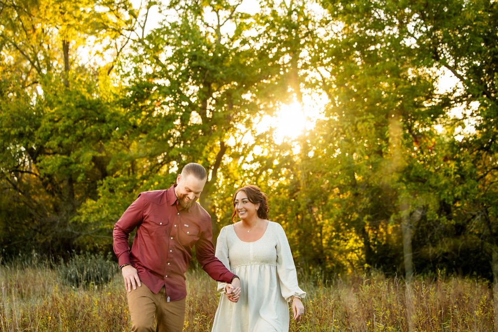 Couple holding hands walking through golden sunlit meadow surrounded by lush green trees during outdoor engagement photo session.
