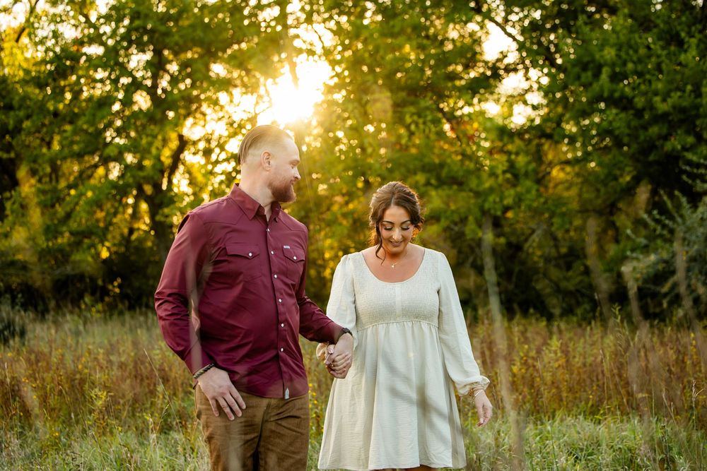 Couple holds hands walking through golden sunlit field at sunset, man in burgundy shirt, woman in white dress.