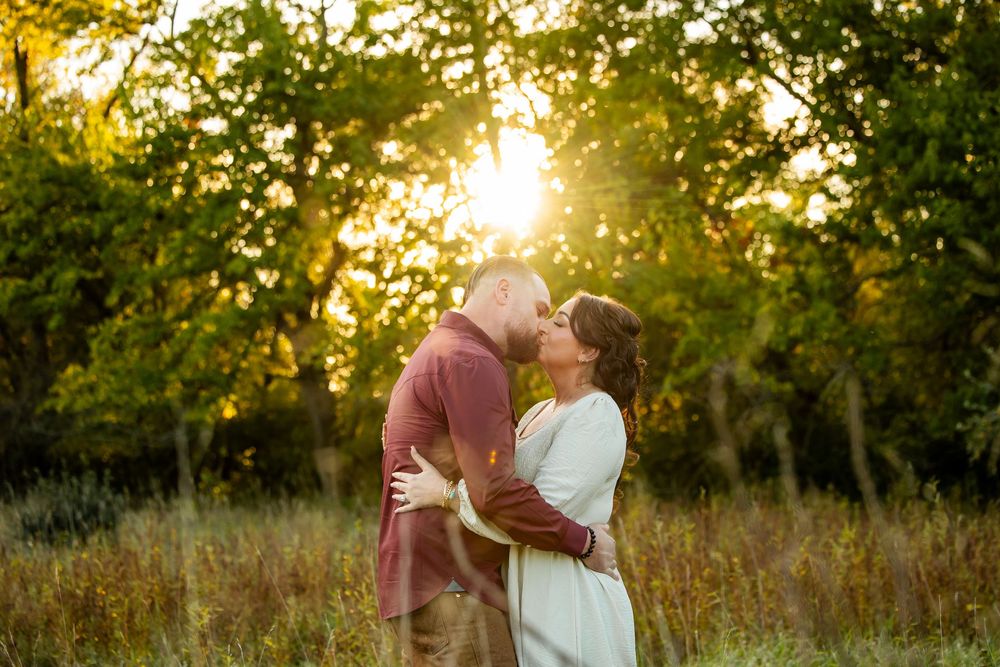 Couple sharing a romantic kiss in a sunlit field surrounded by lush green trees during golden hour.