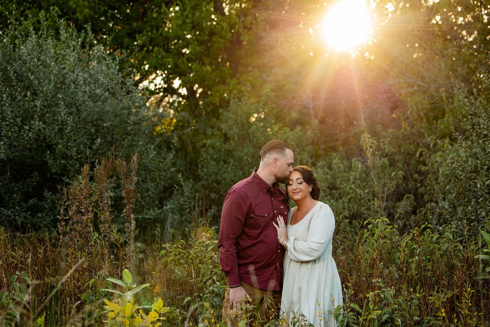 Couple embraces in a sunlit meadow at golden hour, surrounded by lush greenery and warm glowing light.