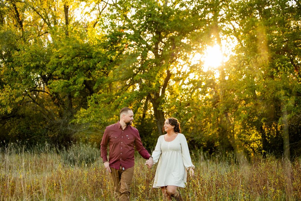 Couple holds hands walking through golden sunlit meadow with lush green trees at sunset during engagement photo session.