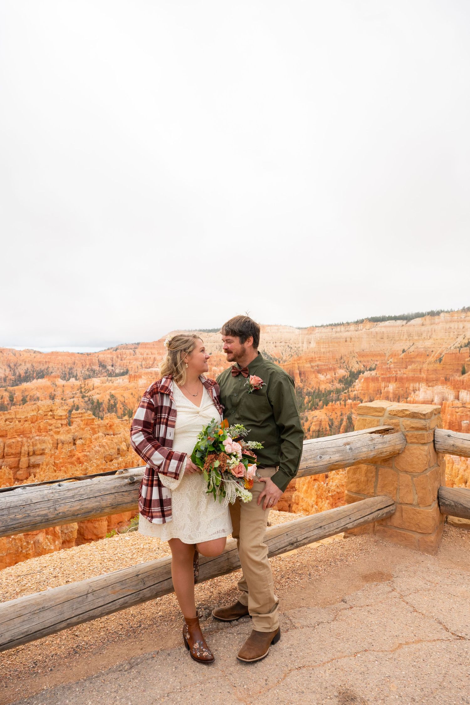 Couple in wedding attire embracing at Bryce Canyon overlook, woman holding bouquet, red rock hoodoos in background.