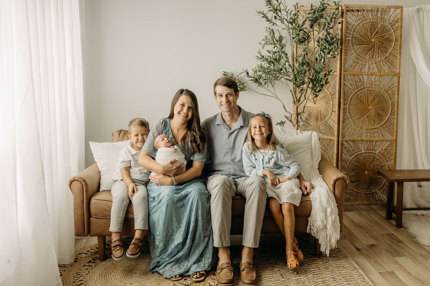 A happy family of five poses on a tan sofa in a bright boho studio, parents holding a newborn and two young children.