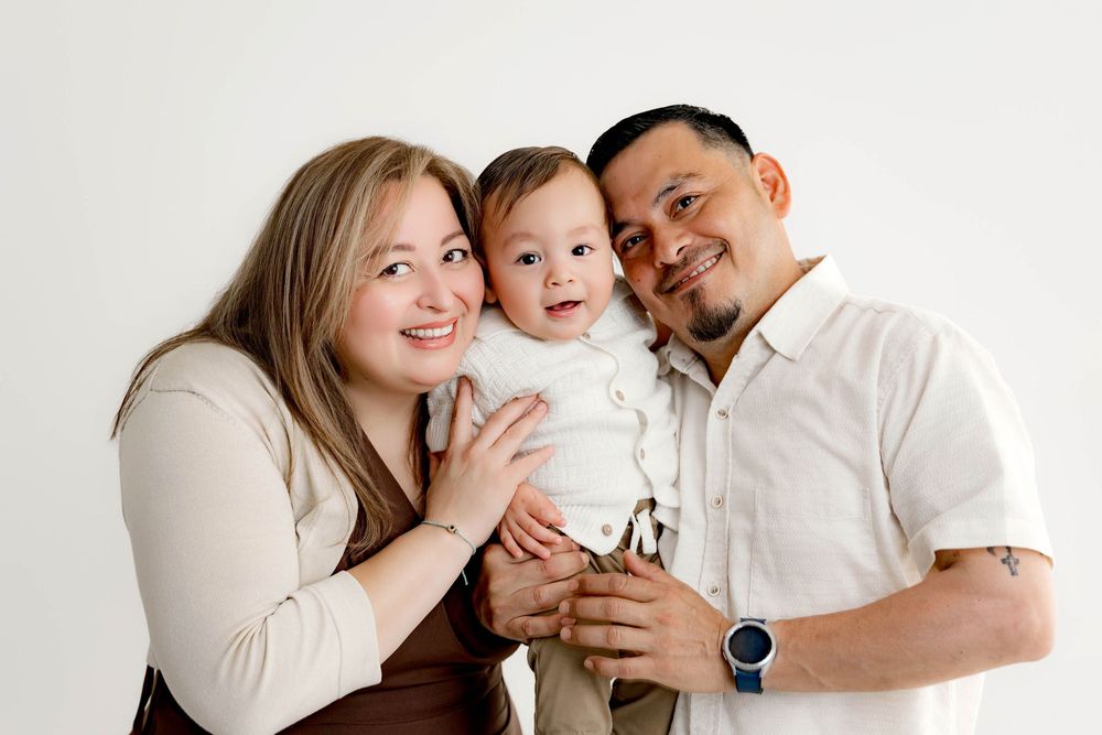Happy family portrait with smiling parents holding their adorable baby in a bright white studio setting.