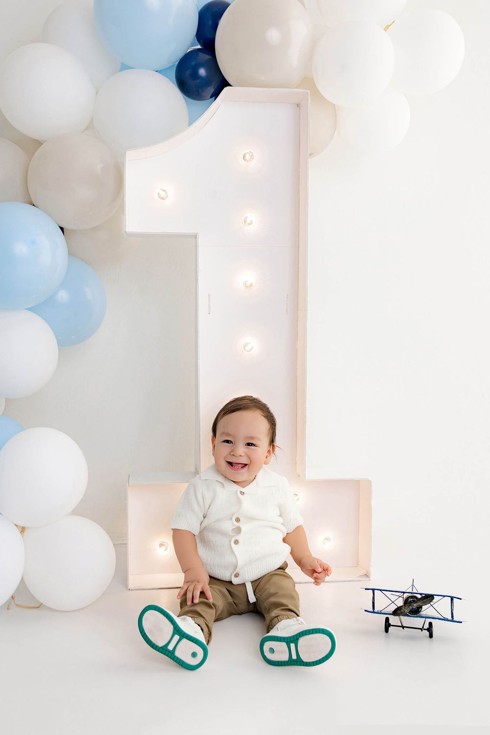 Smiling baby boy sitting in front of a lit number 1 sign with blue and white balloons for his first birthday.
