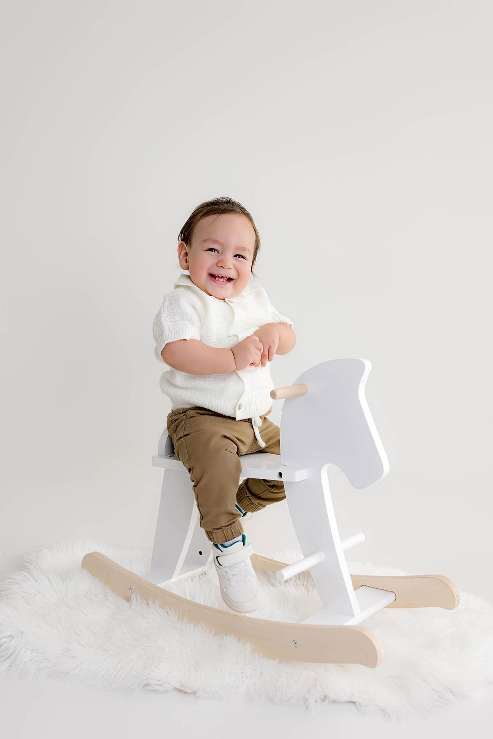 Smiling baby boy sitting on a white wooden rocking horse toy on a fluffy rug against a white background.