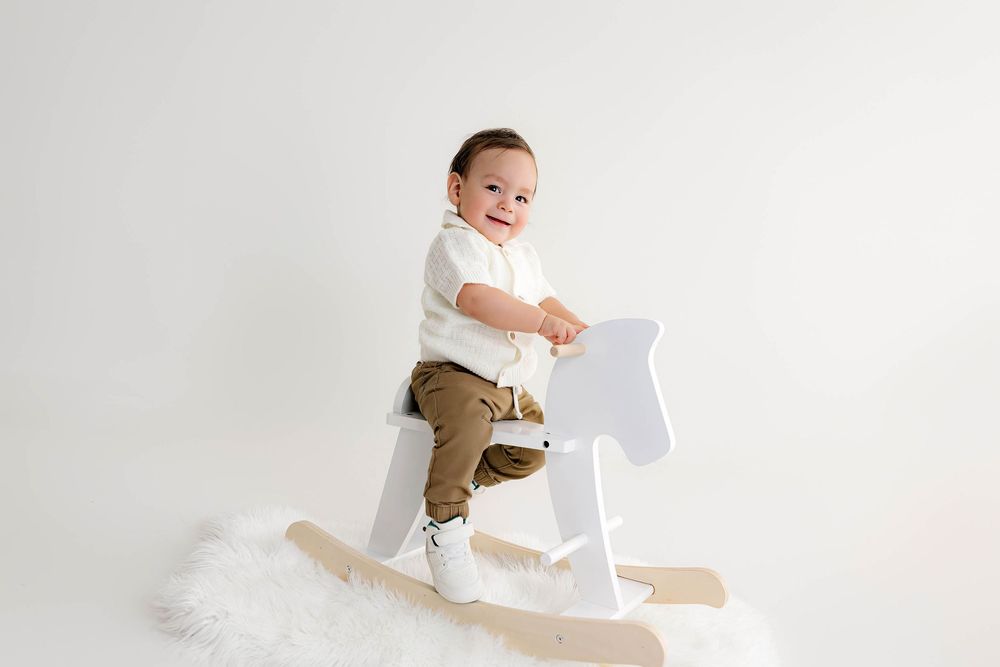 Smiling baby boy in white onesie and brown pants standing on a white wooden rocking horse on a fluffy rug.