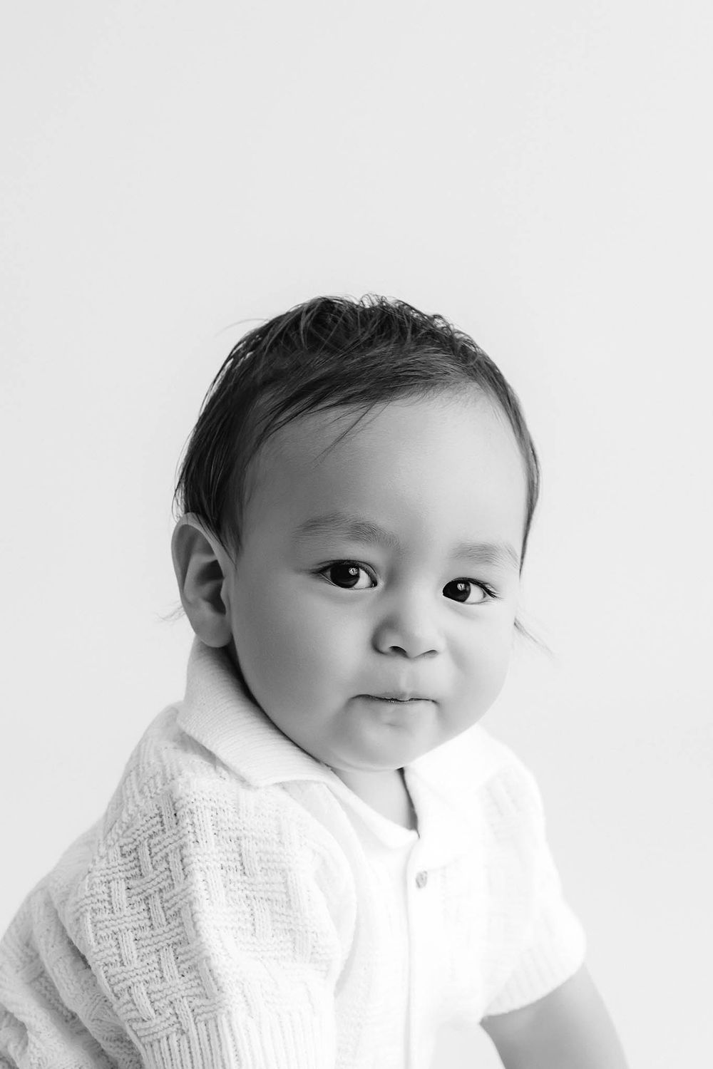 Black and white portrait of an adorable baby in a white knit outfit, gazing softly at the camera.