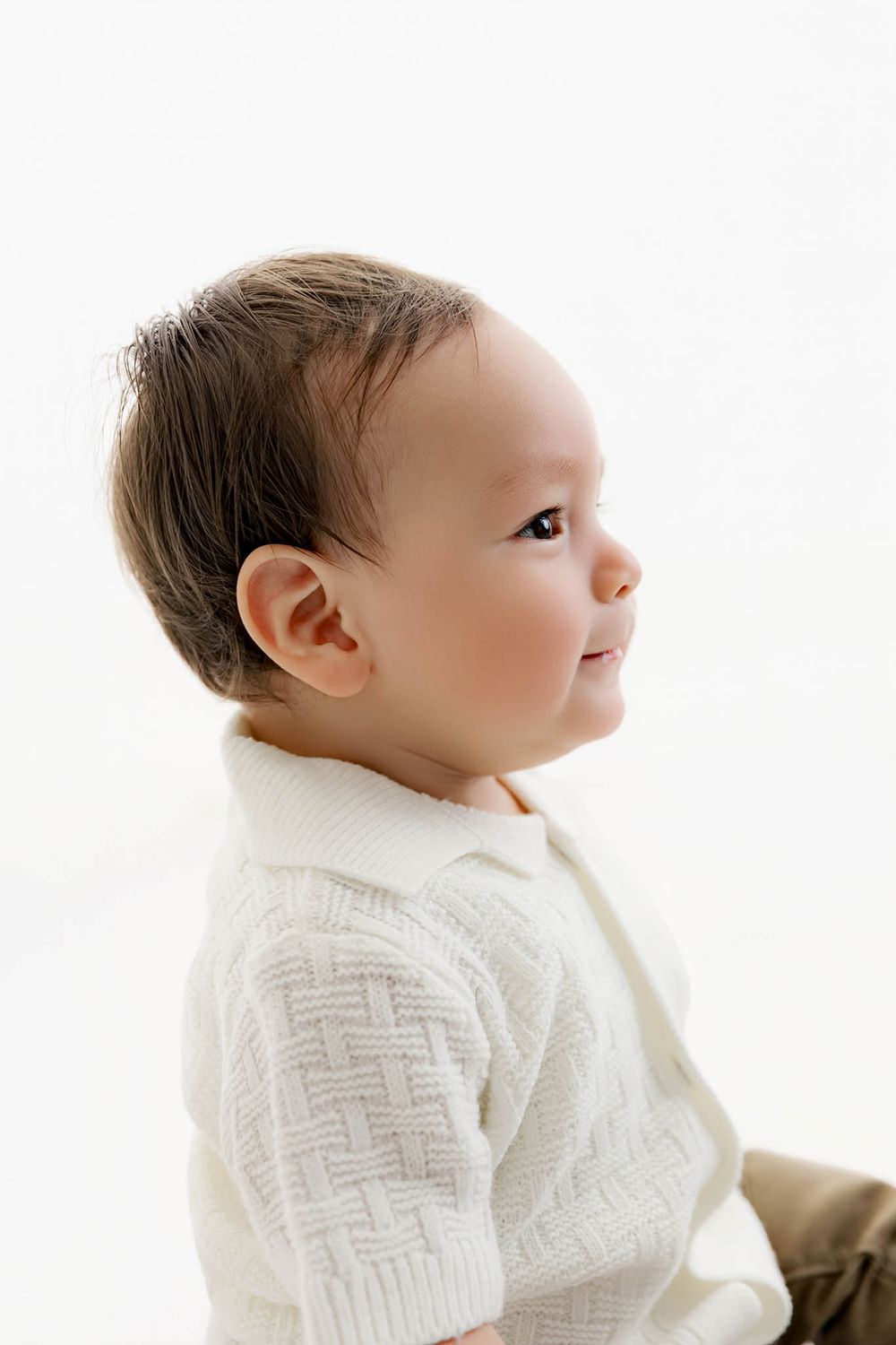 A cute baby in a white knit sweater looks to the side against a bright white background.