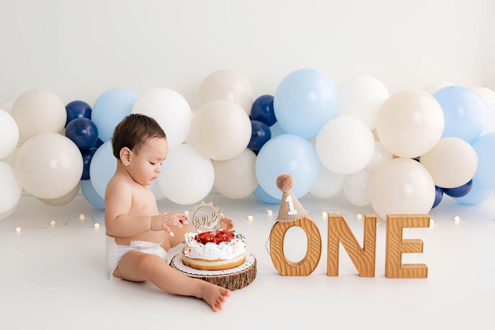 Baby's first birthday cake smash photo with blue and white balloon garland backdrop and wooden ONE letters.